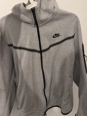 Grå hoodie från Nike med dragkedja - Snygg grå hoodie från Nike med svart logga på bröstet och svarta detaljer. Hoodien har hel dragkedja framtill, svarta linjer över bröstet och en praktisk ficka med dragkedja på ärmen. Perfekt för en sportig och avslappnad stil.