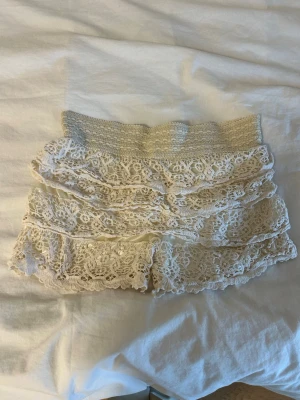 Vita virkade spets-shorts med volanger - Säljer ett par vita virkade shorts med flera lager volanger i spets. De har bred resår i midjan för skön passform och är perfekta för en somrig och bohemisk stil. Materialet är mjukt och luftigt, vilket gör dem riktigt bekväma.