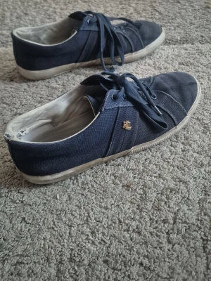 Mörkblå sneakers från Ralph Lauren - Snygga mörkblå sneakers från Ralph Lauren med RL-logga i guld på sidan. Skorna har vita sulor och är tillverkade i canvasmaterial med klassisk snörning. Perfekta för en avslappnad och stilren look.