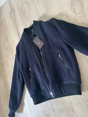 Mörkblå overshirt från Riley - Snygg mörkblå bomberjacka från Riley med dragkedja framtill och två snedställda fickor med silverfärgade dragkedjor. Helt ny med prislapp kvar, nypris 1500kr. Jackan har svarta ribbade muddar vid ärmslut, krage och nederkant. Storlek S, hör av er vid frågor! 