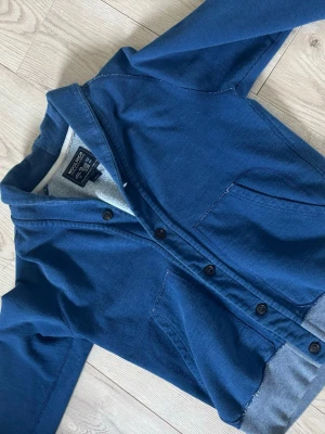 Blå kofta från Woolrich - Snygg blå kofta från Woolrich med knäppning framtill och två stora fickor. Koftan har en avslappnad passform och är tillverkad i mjukt bomullsmaterial. Riktigt snygg nu under våren och 