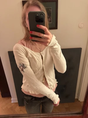 Vit kofta från Odd Molly med brodyr - Söt vit kofta från Odd Molly med broderad detalj på ärmen och dekorativa knappar framtill. Koftan har ribbade muddar och en liten volangkant nedtill. Perfekt för lager på lager och ger en romantisk vibe till din outfit. Den har en liten fläck på baksidan. 