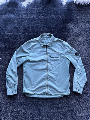 C.P. Company overshirt  - Snygg ljusgrön overshirt från C.P. Company med dragkedja framtill, klassisk krage och bröstficka med lock. Ikonisk linsdetalj på vänster ärm. Tillverkad i ett lätt material som passar perfekt för lager-på-lager. Skick 7/10, har ett hål längst ner på baksidan och nån fläck men övrigt bra skick, kan skicka bild vid intresse. Storlek M. Pris går att diskutera✅