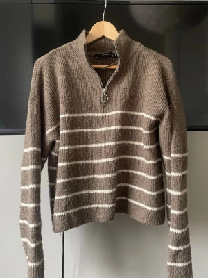 Brun stickad tröja med dragkedja - Mysig brun stickad tröja med vita ränder och halv dragkedja med ringdetalj vid halsen. Tröjan har lång ärm och en avslappnad passform. Super snygg och i väldigt bra skick❣️ 