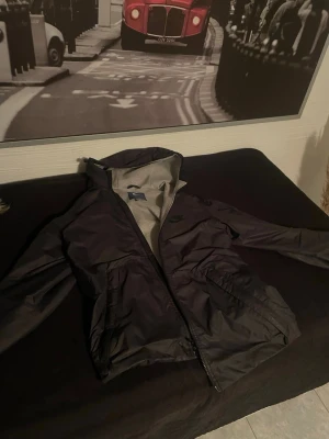 Svart vindjacka från Nike med huva - Nike vindjacka i svart med grått innerfoder och huva. Jackan har dragkedja framtill, två stora fickor och diskret Nike-logga på bröstet. Perfekt för blåsiga dagar och enkel att matcha med streetwear. Materialet är lätt och smidigt.