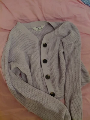 Ljuslila kofta från H&M I storlek 146/152 - Mysig ljuslila kofta från H&M med grovstickad struktur och stora svarta knappar framtill. Koftan har v-ringad hals, långa ärmar och ribbade muddar. Perfekt att slänga över en t-shirt för en avslappnad look. 