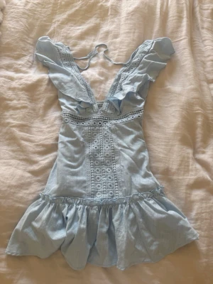 Ljusblå volangklänning med spets - Supersöt ljusblå kortklänning med volanger och spetsdetaljer. Djup v-ringning med knyt i nacken och bred volangkant nedtill. Perfekt för sommaren