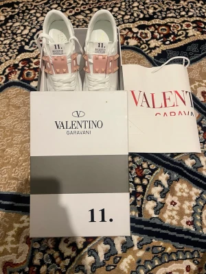 Valentino Garavani vita sneakers - Snygga vita sneakers från Valentino Garavani med rosa band och guldiga nitar över vristen. Skorna har en chunky sula och är tillverkade i läder. Perfekta för dig som vill sticka ut med exklusiva detaljer och modern design.