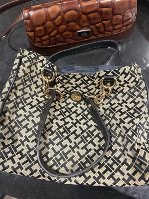 Svart och beige handväska med kedjedetaljer - Snygg handväska i svart och beige med grafiskt mönster och guldfärgade kedjedetaljer. Väskan har svarta handtag, innerfack med dragkedja och rymligt huvudfack. Perfekt för dig som gillar statement-accessoarer och vill ha plats för allt du behöver.