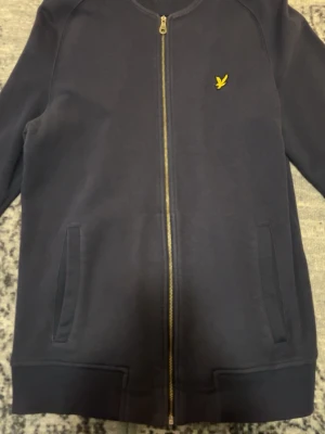 Mörkblå cardiganjacka från Lyle & Scott - Mörkblå cardiganjacka med dragkedja från Lyle & Scott. Jackan har två fickor framtill, ribbade muddar och den klassiska gula loggan på bröstet. Tillverkad i mjukt material som känns bekvämt och stilrent.
