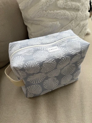 Blå necessär med snäckmönster från Emelo - Necessär i ljusblått tyg med vita snäckskal som mönster. Rektangulär form med dragkedja upptill och ett praktiskt handtag på sidan. Märkt med Emelo på framsidan. Perfekt för smink eller småprylar.