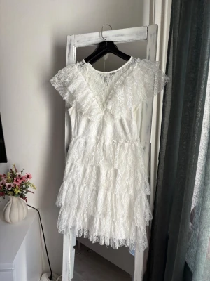Vit studentklänning  - Superfin vit kortklänning i spets med flera lager volanger och v-ringad hals. Klänningen har korta ärmar med volangdetaljer och en romantisk känsla. Perfekt för studenten! Endast använd på studenten.