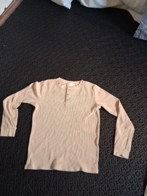 Beige ribbad långärmad tröja H&M - Säljer en beige ribbad långärmad tröja från H&M med knappar vid halsen. Tröjan har en klassisk rund halsringning och är gjord i mjukt bomullsmaterial. Perfekt för lager på lager och enkel att matcha med olika outfits.