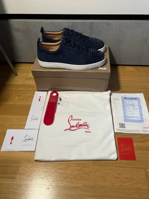 Mörkblå Christian Louboutin sneakers - Snygga mörkblå sneakers från Christian Louboutin med ikoniska röda sulor och coola nitar framtill. Skorna är låga och har ovandel i mocka med matchande blå skosnören. Perfekt för dig som vill sticka ut med exklusiv streetstyle. Helt nya, oanvända