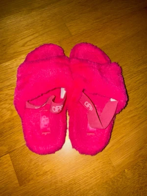 Fluffiga rosa tofflor med band - Säljer ett par riktigt fluffiga och mjuka tofflor i starkt rosa färg. De har öppna tår och breda band över foten med textdetalj. Sulan är svart och räfflad för bättre grepp. Perfekta för att chilla hemma och hålla fötterna varma.
