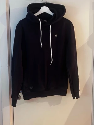 Mörkblå morris hoodie - Mörkblå morris hoodie med vit broderad lilja på bröstet och vita dragsnören i huvan. Klassisk modell med ribbade muddar och mjukt material, perfekt för en avslappnad men även stilren stil.