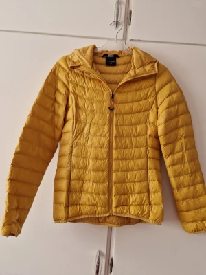 Gul pufferjacka med huva från H&M - Snygg gul pufferjacka från Stadium med huva och dragkedja framtill. Jackan har quiltad design med horisontella sömmar och två sidofickor. Perfekt för kalla vinterdagar och enkel att matcha med olika outfits.