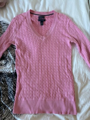 Rosa kabelstickad tröja - Snygg rosa kabelstickad tröja från Tommy Hilfiger med V-ringning och långa ärmar. Tröjan har ribbade muddar vid ärmslut och nederkant, och är gjord i mjukt material som sitter skönt på kroppen.