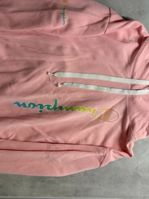 Rosa hoodie från Champion - Snygg rosa hoodie från Champion med färgglad logga framtill. Hoodien har huva med vita dragsnören och är tillverkad i mjukt material, perfekt för en avslappnad stil.