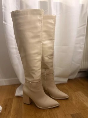 Beiga höga stövlar med klack - Snygga knähöga stövlar i beige konstläder med bred klack och spetsig tå. Dragkedja på insidan för enkel påtagning. Perfekta för att lyfta din outfit med en trendig och stilren look. Dom är i storlek 38 tror jag. 