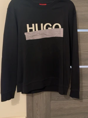 Svart sweatshirt från HUGO - Äkta Cool svart sweatshirt från HUGO med stor vit logga och grå detalj framtill. Tröjan har rund halsringning, långa ärmar och ribbade muddar. Perfekt för en avslappnad och trendig look.
