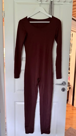 Mörkbrun långärmad jumpsuit - En tight, mörkbrun jumpsuit med långa ärmar och rund halsringning. Modellen är figurnära och hel i benen, perfekt för en trendig och minimalistisk look. Materialet ser ut att vara mjukt och stretchigt, vilket gör den bekväm att bära hela dagen.