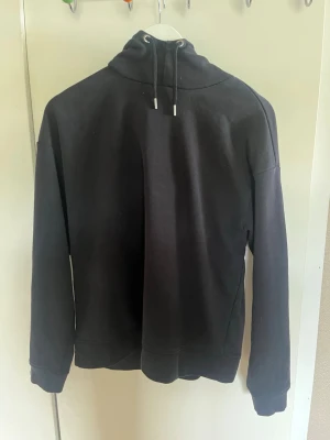 Svart hoodie med dragsko - En stilren svart hoodie med huva och dragsko. Hoodien har långa ärmar och ribbade muddar vid ärmslut och nederkant. Perfekt för en avslappnad look och enkel att matcha med andra plagg.