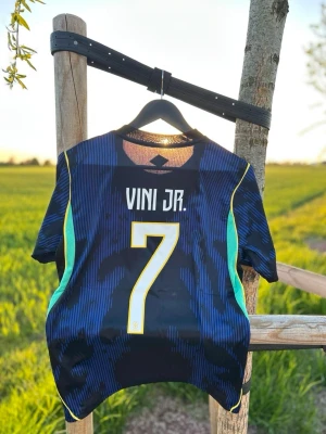 Brasilien Vini Jr. matchtröja #7 - Snygg mörkblå Brasilien matchtröja med Vini Jr. och nummer 7 på ryggen. Tröjan har gula och turkosa detaljer, CBF-logga och Jordan-logga på bröstet. Tillverkad i lätt och ventilerande material, perfekt för fotboll eller träning.