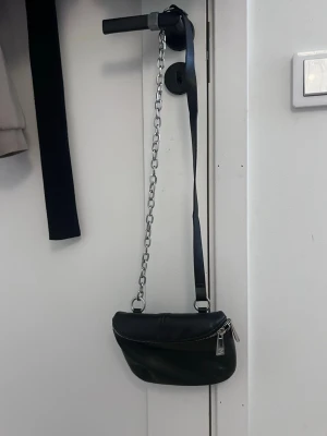 Svart axelremsväska med kedjedetalj - Cool svart handväska i slätt konstläder med silverfärgad kedja och justerbar axelrem. Väskan har en rundad form, dragkedja upptill och en praktisk ficka på baksidan. Perfekt för att lyfta din outfit med en edgy touch.
