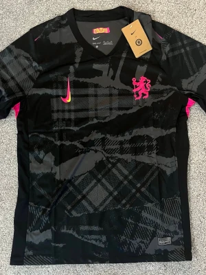 Svart Nike Chelsea matchtröja - Snygg svart träningströja från Nike med BLUFS-logga och rosa detaljer. Tröjan har ett unikt rutmönster, rosa lejonemblem och Nike swoosh i rosa och gult. Tillverkad i lätt och ventilerande material, perfekt för träning eller sport. Bara skicka meddelande vid frågor👍 Sorry för sista bilden