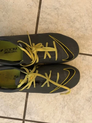 Nike Mercurial fotbollsskor grå/gul - Säljer ett par Nike Mercurial fotbollsskor i grått med gula detaljer och gula skosnören. Skorna har en smal och aerodynamisk form, med gula ränder och logga på sidan. Yttermaterialet är syntetiskt och sulan har fasta dobbar för spel på gräs.