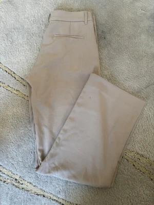 Beige kostymbyxa med raka ben från Gina tricot - Stilrena beige kostymbyxor med raka ben och klassisk passform. Byxorna har hällor för bälte, sidfickor och en bakficka med passpoal. Perfekta för en dressad look. Materialet känns mjukt och lätt, troligtvis bomulls- eller polyestermix.