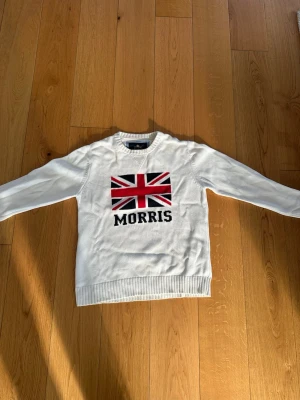 Vit stickad tröja Morris Union Jack - Vit stickad tröja från Morris med Union Jack-flagga i rött, svart och vitt på bröstet samt texten 'MORRIS' under. Tröjan har rund halsringning och ribbstickade muddar vid ärmslut och nederkant. Perfekt för en stilren och klassisk look. XL men passar L bättre