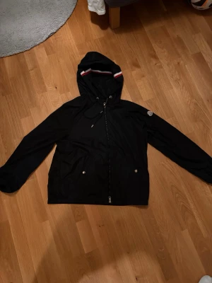 Svart Moncler vindjacka med huva - Svart vindjacka från Moncler med huva och vit-röd rand på huvan. Jackan har dragkedja framtill, två fickor med tryckknappar och Moncler-logga på ärmen. Lätt och smidig modell, perfekt för blåsiga dagar.