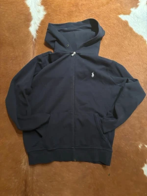 Mörkblå hoodie från Polo Ralph Lauren - Mörkblå hoodie med dragkedja från Polo Ralph Lauren. Klassisk design med huva, fickor framtill och den ikoniska vita logon broderad på bröstet. Perfekt för en chill och stilren look.