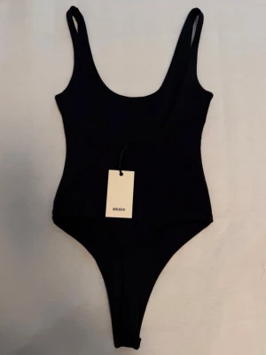 Svart body från Bik Bok - Stilren svart body från Bik Bok med djup rundad urringning och smala axelband. Modellen har högt skurna ben och är tillverkad i ett stretchigt material som sitter tajt mot kroppen. Perfekt för att skapa en snygg siluett under kläder. Söt med jeans och kjolar 🤍