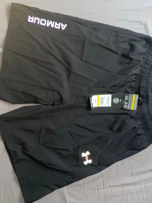 Svarta träningsshorts Under Armour - Svarta träningsshorts från Under Armour med elastisk midja, reflekterande logga framtill och texten 'ARMOUR' på ena benet. Lätta och luftiga, perfekta för gym eller löpning. Snygga detaljer baktill med reflekterande streck. Materialet känns sportigt och snabbtorkande.