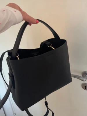 Svart handväska arket - Snygg svart handväska med minimalistisk och stilren form. Väskan har både handtag och axelrem, samt guldfärgade metalldetaljer. Tillverkad i slätt konstläder som ger en modern look. Perfekt för dig som gillar enkel och trendig stil. Den stora modellen 