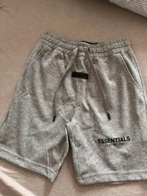 Grå Essentials Fear of God shorts - Säljer ett par grå shorts från Essentials Fear of God med snörning i midjan och två sidofickor. Klassisk logga tryckt på vänster ben. Sköna och stilrena shorts i mjuk bomullsblandning, perfekta för sommaren.