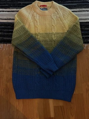 Ombre stickad tröja från Carli - Stickad tröja i 100% ull från Carli med snygg ombre-effekt i beige, grönt och blått. Tröjan har grovstickat mönster med flätor framtill, rund halsringning och ribbade muddar. Perfekt för dig som gillar färg och vill ha något unikt i garderoben.