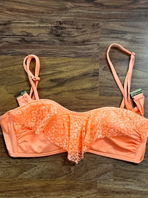 Orange bikiniöverdel med spetsvolang - Snygg orange bikiniöverdel med volang i spets framtill. Justerbara axelband och dekorativ spetsdetalj ger en trendig look. Perfekt för strandhäng och poolparty. Materialet känns mjukt och följsamt mot huden.