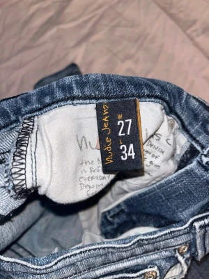 Blå jeans från Nudie Jeans, W27 L34 - Snygga blå jeans från Nudie Jeans med klassisk femficksdesign och mörkblå broderade detaljer på bakfickorna. Jeansen har en något tvättad look och är tillverkade i bomull. Perfekta för en avslappnad och tre