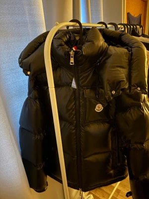 Svart dunjacka från Moncler - Snygg svart dunjacka från Moncler med glansig finish, huva och dragkedja framtill. Jackan har en bröstficka med lock och Moncler-logga, samt flera praktiska fickor. Perfekt för kalla vinterdagar och riktigt trendig stil.