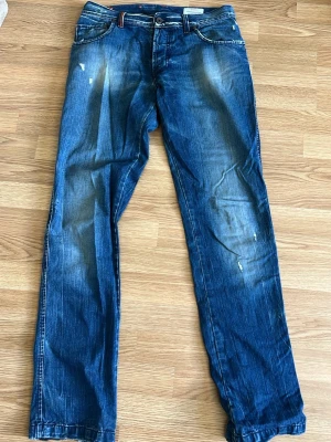Blå raka jeans från Boss Orange - Säljer ett par blå jeans från Boss Orange med raka ben och slitna detaljer framtill. Jeansen har klassisk femficksmodell, knappgylf och tydliga slitningar för en cool, avslappnad look. Perfekta för dig som gillar streetstyle och vintagekänsla.
