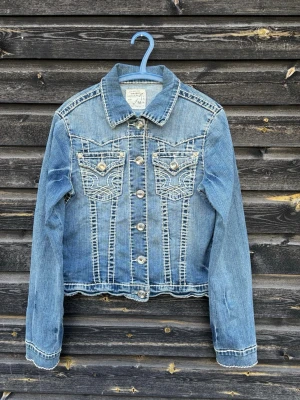 Blå jeansjacka från True Religion - Snygg blå jeansjacka från True Religion med kontrastsömmar och två bröstfickor med dekorativa knappar. Klassisk krage och knappstängning framtill. Jackan har en figurnära passform och coola detaljer som ger en trendig look.