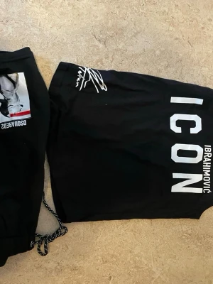 Svart ICON t-shirt från Dsquared2 - Snygg svart t-shirt från Dsquared2 med stor vit text 'ICON IBRAHIMOVIC' på ryggen och signaturdetalj vid axeln. Framsidan har ett porträtt och citat i vitt. Perfekt för dig som gillar streetwear och vill sticka ut.