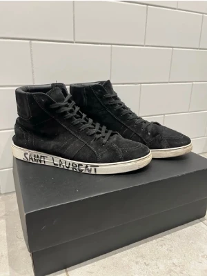 Svarta höga sneakers från Saint Laurent - Svarta höga sneakers från Saint Laurent i mocka med klassisk siluett och svarta skosnören. Vit sula med handskriven logga på sidan ger en cool och unik touch. Perfekta för dig som gillar streetstyle och exklusiva märken.passar som 43/44
