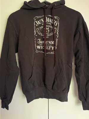 Jack Daniel's svart hoodie - Svart hoodie med stor Jack Daniel's Tennessee Whiskey-logga i vitt tryck både fram och bak. Klassisk känguruficka, huva med dragsko och ribbade muddar. Perfekt för dig som gillar ikoniska tryck och vill ha en avslappnad streetwear-look.