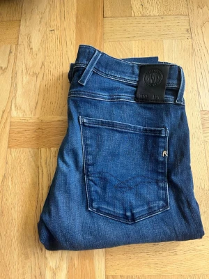 Replay Anbass - Snygga blå slim fit jeans från Replay med klassisk femficksdesign och diskret slitning. Jeansen har en mörkblå tvätt, raka ben och detaljer som logotyp på bakfickan och läderpatch i midjan. Perfekta för dig som gillar stilrena och moderna jeans. MODELL: Anbass (slim fit) W32 L32