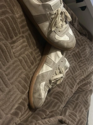 Maison marginal skor  - Snygga sneakers i vitt och beige med klassisk design. Skorna har ovandel i läder och mocka, samt en brun gummisula. Perfekta för dig som gillar stilrena och tidlösa sneakers med retrovibbar. Skosnören i matchande färg och låg profil. Kan tvätta dem innan ni får dem! 